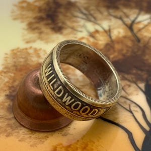 May include: A vintage-style ring with the word "WILDWOOD" engraved in bold letters. The ring has a textured band and a bronze finish. The ring is resting on a copper-colored thimble.