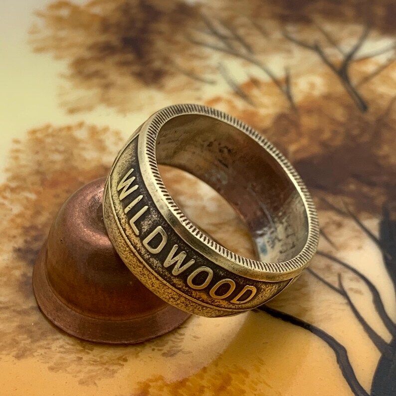 May include: A vintage-style ring with the word "WILDWOOD" engraved in bold letters. The ring has a textured band and a bronze finish. The ring is resting on a copper-colored thimble.