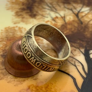 May include: A vintage-style brass ring with the word "WILDWOOD" engraved around the band. The ring has a worn, antique appearance. The ring is resting on a copper-colored object.