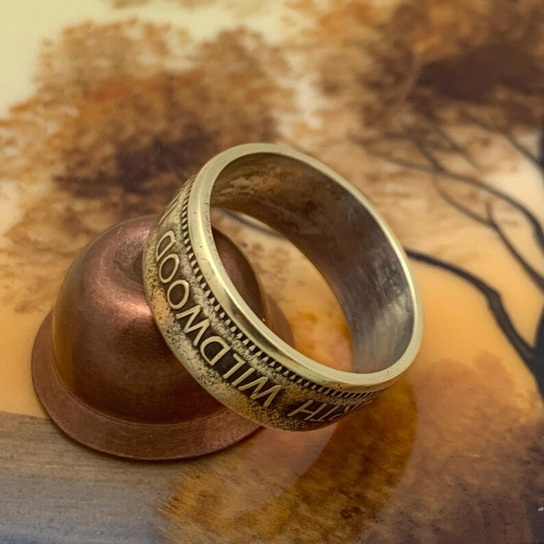 May include: A close-up of a silver ring with the inscription "WILDWOOD" around the band. The ring is resting on a small, copper-colored object. The background is a blurred, warm-toned pattern.