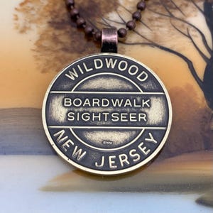 May include: A brass-colored pendant necklace with a round charm that says "WILDWOOD BOARDWALK SIGHTSEER NEW JERSEY" in black lettering. The charm has a worn, antique look.