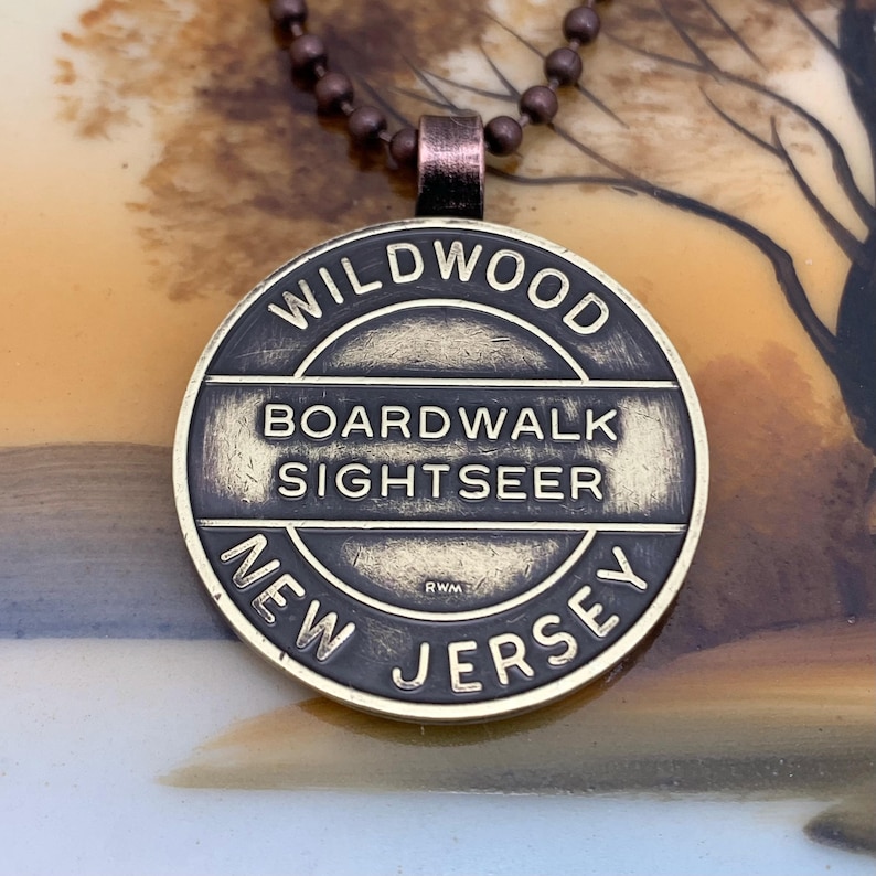 May include: A brass-colored pendant necklace with a round charm that says "WILDWOOD BOARDWALK SIGHTSEER NEW JERSEY" in black lettering. The charm has a worn, antique look.