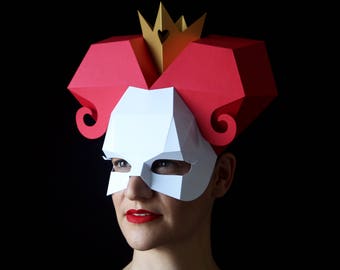May include: A white paper mask with a red heart on the forehead and a red crown. The mask has eye holes and a red paper design around the eyes. The mask is worn by a person with red hair.