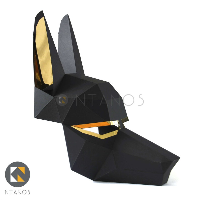 May include: A black geometric Anubis mask with gold foil accents. The mask is made of folded paper and features sharp angles and a stylized dog-like face. The ears and eye areas are lined with gold foil. The mask is a decorative item.