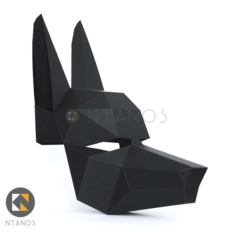 May include: A black geometric papercraft mask in the shape of a wolf or dog head. The mask has sharp angles and a polygonal design. The mask is made of black paper and is displayed against a white background. The logo "NTANOS" is visible.