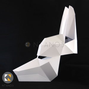 May include: A white, geometric paper mask in the shape of a canine head, with pointed ears and a triangular snout. The mask is constructed from folded paper, creating a faceted, three-dimensional effect. The logo "NTANOS" is visible in the lower left corner.