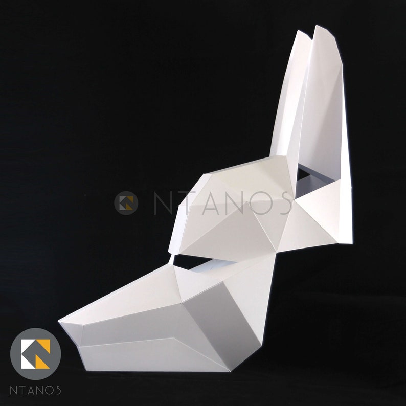May include: A white, geometric paper mask in the shape of a canine head, with pointed ears and a triangular snout. The mask is constructed from folded paper, creating a faceted, three-dimensional effect. The logo "NTANOS" is visible in the lower left corner.