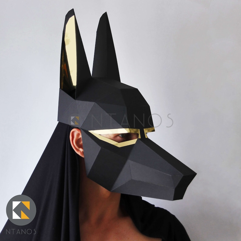 May include: A black geometric mask with gold accents, resembling the head of a jackal. The mask has a stylized, angular design and is worn by a person. The mask is a unique costume piece.