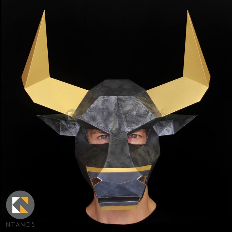 May include: A black and gold bull mask with a textured surface. The mask has a pointed nose and large, pointed ears. The eyes are cut out and the mask is worn by a person.