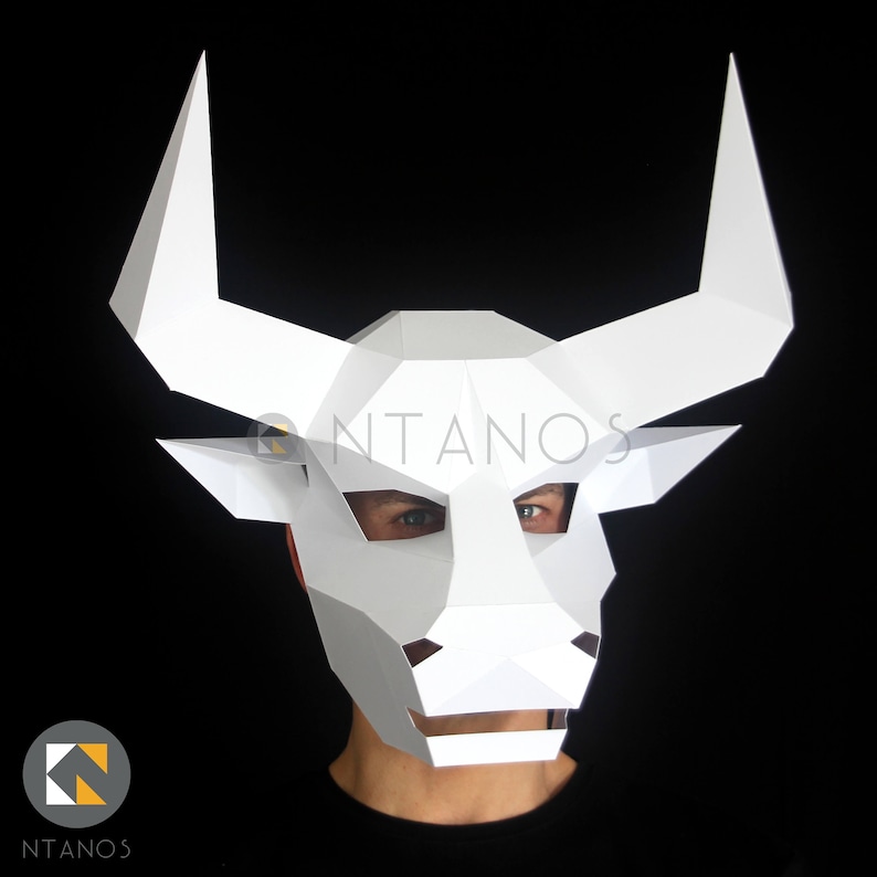 May include: White papercraft bull mask with large horns. The mask is worn by a person with a black shirt. The mask has a geometric design and the word "NTANOS" is visible on the mask.