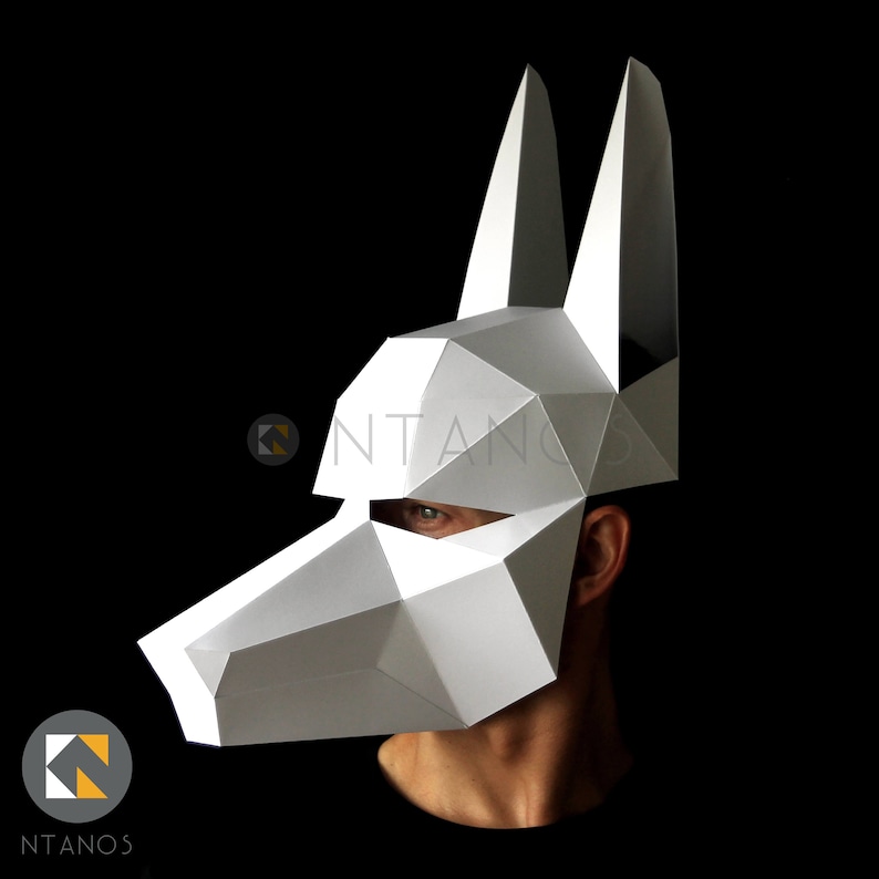May include: A white geometric wolf mask with pointed ears and a long snout. The mask is made of folded paper or cardstock, with visible edges and angles. The mask covers the face, with the wearer's eye visible. The background is black.