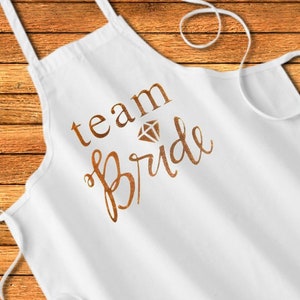 May include: White apron with the text "team bride" in gold lettering with a diamond graphic above the word "bride".