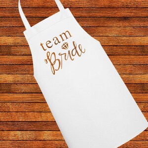 May include: White apron with the text "team bride" in gold lettering and a diamond graphic.