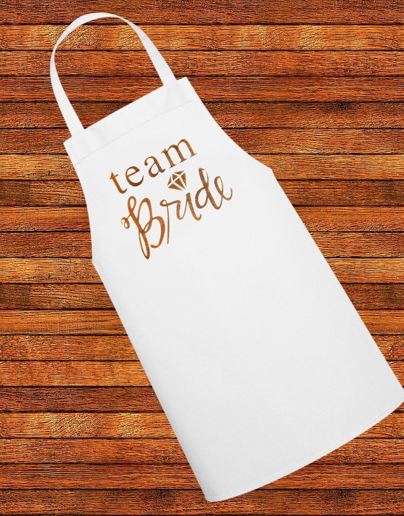 May include: White apron with the text "team bride" in gold lettering and a diamond graphic.
