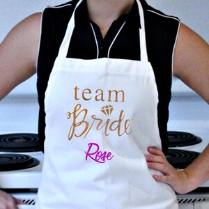 May include: White apron with the text "team bride" in gold script with a diamond graphic above the text. The name "Rose" is written in pink script below the text.
