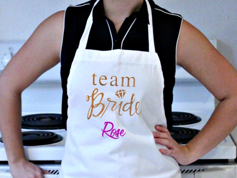 May include: White apron with the text "team bride" in gold script with a diamond graphic above the text. The name "Rose" is written in pink script below the text.
