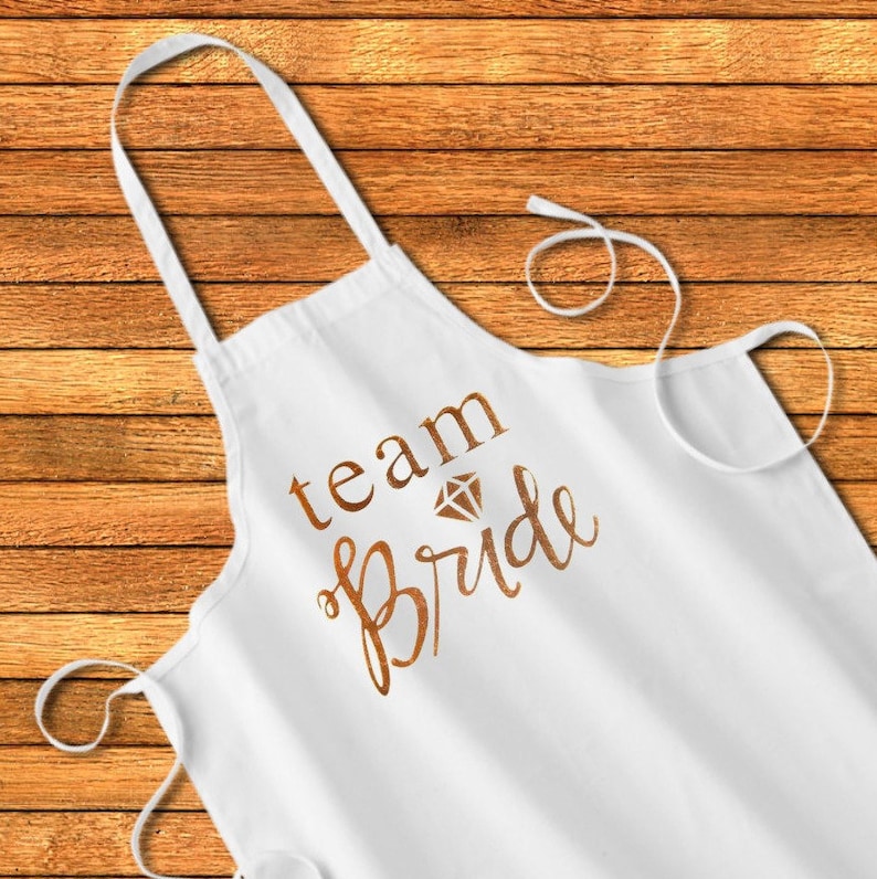 May include: White apron with the text "team bride" in gold lettering with a diamond graphic above the word "bride".