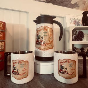 May include: A white glittery travel mug and two matching mugs with black handles. Each features a vintage-style label with a cartoon character and the text "Whoville Fresh Hot Delicious Hot Cocoa". A black and brown thermos is also in the image.