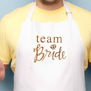 May include: White apron with the text "team bride" in brown lettering with a diamond graphic above the word "bride". A person is wearing the apron and holding a fork and a black spatula.