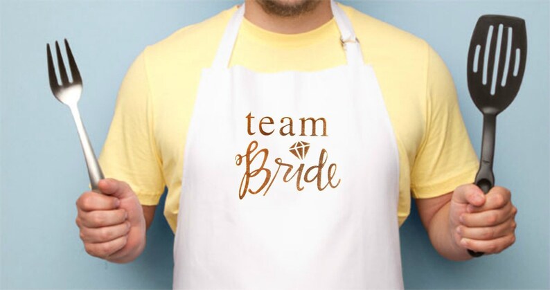 May include: White apron with the text "team bride" in brown lettering with a diamond graphic above the word "bride". A person is wearing the apron and holding a fork and a black spatula.