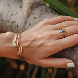 Pode incluir: Um conjunto de pulseira e anel dourados. A pulseira tem um design curvo e o anel apresenta um padrão floral com uma pedra central azul. A mão está apoiada num galho de árvore, com uma manga estampada visível.