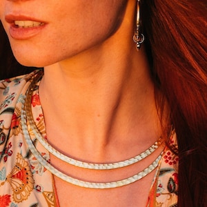 May include: A light beige fedora hat with a wide brim. The person is wearing a floral patterned top and a layered gold necklace. The person has long red hair and is wearing hoop earrings.
