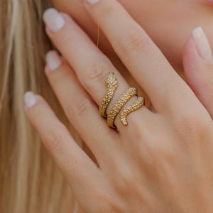 Könnte beinhalten: Ein goldfarbener Schlangenring umschlingt einen Finger. Der Ring hat eine strukturierte Oberfläche, wobei sich Kopf und Schwanz der Schlange treffen. Ein Perlenarmband mit geometrischem Muster und ein weißes Perlenarmband sind ebenfalls zu sehen.