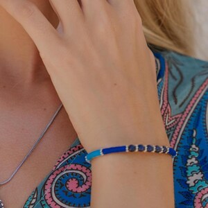 Op de afbeelding: Een zilveren ketting met een ovale hanger met een donkere steen, een bijpassende ring en een blauw en zilveren armband. De vrouw draagt een blauw-roze paisley top en een zwarte oorbel met een vlinderontwerp.
