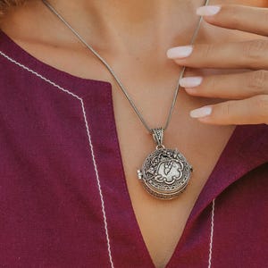 Pode incluir: Um pendente de medalhão circular em tom prateado numa corrente fina, com padrões florais e de volutas em relevo. O conjunto de joalharia inclui brincos pendentes a condizer com um design ornamentado e um anel largo ajustável.