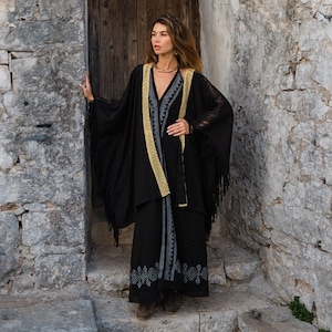 Op de afbeelding: Een zwart ensemble met een lange, vloeiende jurk en een bijpassende sjaal met franjes. De jurk heeft een V-hals en geborduurde details. Een goudkleurige bies accentueert de sjaal en de jurk. De outfit wordt gedragen met een gouden ketting.