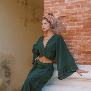 Peut inclure: Ensemble deux pièces vert foncé composé d'une jupe portefeuille et d'un haut noué à manches évasées. La jupe et les manches présentent un motif brodé noir. Un foulard à motifs et des bijoux complètent le look.