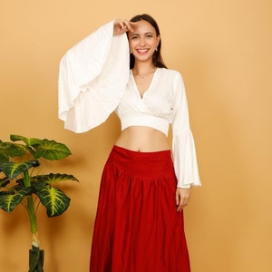 Könnte beinhalten: Eine Frau trägt ein weißes Crop-Top mit ausgestellten Ärmeln und einen roten Maxirock. Das Oberteil hat einen V-Ausschnitt und eine taillierte Taille. Der Rock ist einfarbig rot und reicht bis zu den Knöcheln. Das Outfit wird barfuß und vor neutralem Hintergrund getragen.