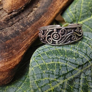 Può includere: Un anello argentato con un design a spirale intricato. L'anello presenta una serie di spirali e curve, creando un motivo dettagliato. L'anello è ambientato su uno sfondo di foglie verdi e un pezzo di legno stagionato.