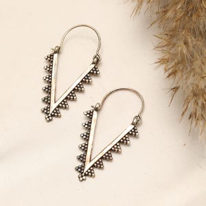 May include: A pair of silver-toned hoop earrings featuring a V-shaped design. Each earring has a pointed bottom and is decorated with small, raised metal beads along the outer edges, suspended from a thin, curved wire closure mechanism.