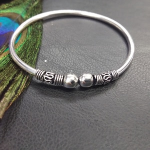 May include: A silver-colored bangle bracelet with decorative detailing. The bracelet features two silver beads and ornate patterns near the center. The design includes a series of lines and a stylized swirl pattern. The bracelet is displayed on a dark surface.