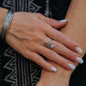 May include: A silver cuff bracelet and ring. The cuff has a textured pattern, and the ring features an open design with a small, pale stone. The jewellery is displayed on an arm, with a black dress visible in the background.