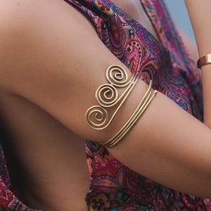Pode incluir: Uma mulher usando joias de ouro, incluindo um colar, pulseira e bracelete de braço. O bracelete apresenta um design em espiral. Ela também está usando um vestido com estampa paisley colorido com costas baixas e alças amarradas.
