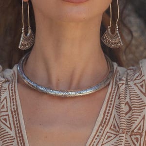 Könnte beinhalten: Eine Frau trägt eine silberne Halskette, Ohrringe und ein Armband. Die Halskette ist ein dickes Band mit strukturiertem Design. Die Ohrringe sind lang, mit einem fächerförmigen Anhänger. Das Armband ist ein einfacher Armreif. Sie trägt ein cremefarbenes Oberteil mit geometrischem Muster.