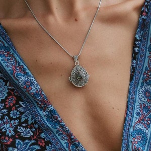 Pode incluir: Um colar de prata com um pingente em forma de gota, um anel a condizer e uma pulseira de prata com um nó. As joias são exibidas sobre um vestido azul com padrão floral.