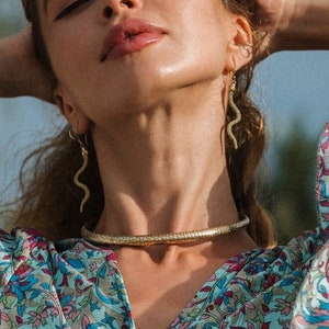 May include: A woman wearing a floral print wrap top with puffed sleeves, gold jewelry, and a gold choker necklace. The earrings are in the shape of a snake. A gold bracelet is on the left wrist.