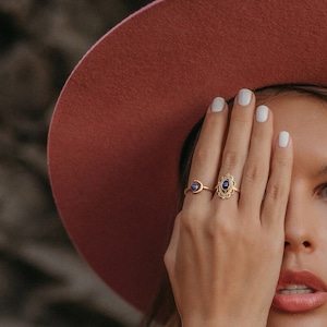 Op de afbeelding: Een vrouw draagt een stofroze breedgerande hoed, gouden sieraden en een roze paisley jurk. De sieraden omvatten een ring met een blauwe steen, een gouden manchetarmband en een gouden choker-ketting. De hand van de vrouw bedekt de helft van haar gezicht.