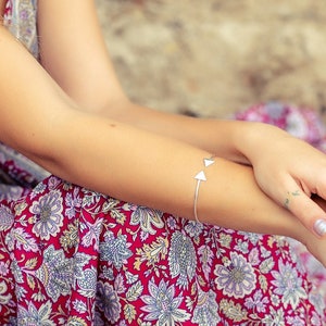 Può includere: Un vestito rosso con stampa floreale con un motivo floreale bianco e blu. Sono visibili un braccialetto d'argento con due ciondoli a triangolo e un anello d'argento con un design circolare. La persona è seduta sulla sabbia.