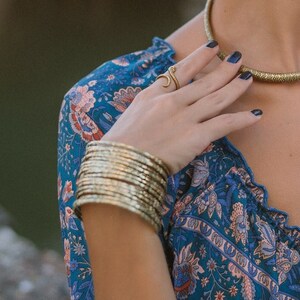 May include: A woman wearing gold jewelry, including a necklace, earrings, ring, and multiple bangles. The jewelry complements a blue floral print dress and a patterned headscarf. The jewelry has a textured, metallic finish.