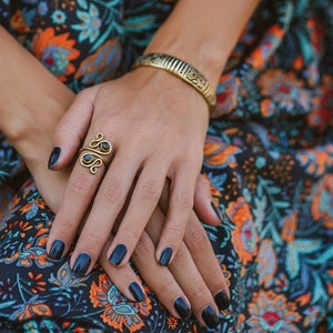 Könnte beinhalten: Ein goldfarbener Ring mit einem Wirbeldesign und ein passendes Armband werden gezeigt. Die Hände sind mit dunkelblauem Nagellack geschmückt, vor dem Hintergrund eines floral gemusterten Kleides in Marineblau, Orange und Türkis.