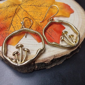 Könnte beinhalten: Goldfarbene Creolen mit einem Pilzdesign. Die Ohrringe haben ein strukturiertes, schlangenartiges Band, das den Reifen bildet, mit kleinen Pilzfiguren im Inneren. Die Ohrringe werden auf einer Holzoberfläche mit Herbstblättern präsentiert.