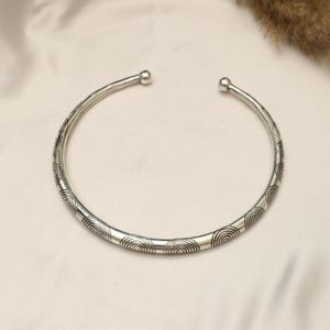 Op de afbeelding: Een zilverkleurige metalen choker-halsketting met een cirkelvormig ontwerp met open uiteinden. Het oppervlak van de band is versierd met een herhalend patroon van gegraveerde concentrische halve cirkels en kleine bolletjes aan de uiteinden.