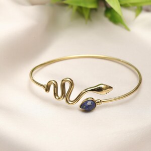 Könnte beinhalten: Goldfarbenes Schlangenarmband mit blauem Edelstein-Akzent. Das Armband hat ein gewundenes Schlangendesign mit detailliertem Kopf und einem tropfenförmigen blauen Stein. Das Armband ist vor einem weißen Hintergrund platziert.