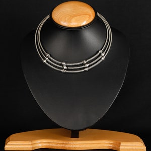 May include: A silver-toned necklace with three strands, each featuring a series of small, round beads and coiled metal accents. The necklace is displayed on a black jewelry stand with a wooden base and top.