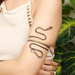 Op de afbeelding: Een zilverkleurige armmanchet in de vorm van een slang. De slang wikkelt zich om de arm, met de kop aan de bovenkant en het lichaam dat zich om de arm wikkelt. De manchet wordt op de bovenarm gedragen.