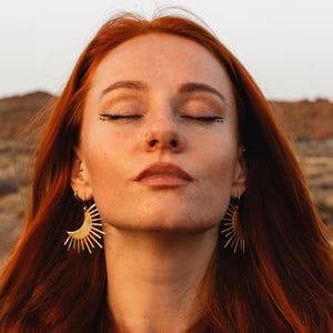 May include: A woman with red hair wearing gold sunburst earrings. The earrings are a crescent shape with radiating lines. The woman has closed eyes and a nose ring. The background is a blurred landscape.